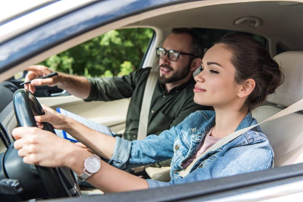 A woman in a denim jacket is driving a car, focused on the road. A man in glasses, sitting beside her, is pointing at the dashboard and holding a clipboard, appearing to give instructions. Both are wearing seat belts.