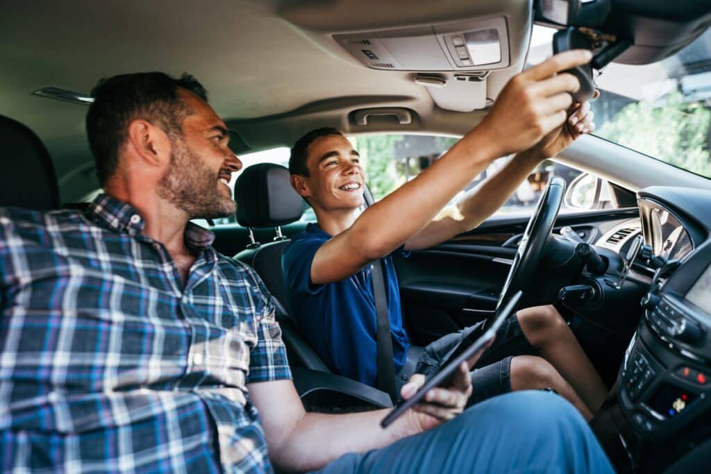 A smiling teenage boy is adjusting the rearview mirror in a car while an adult man, sitting in the front passenger seat, looks at him proudly. Both appear to be sharing a joyful moment inside a parked vehicle.