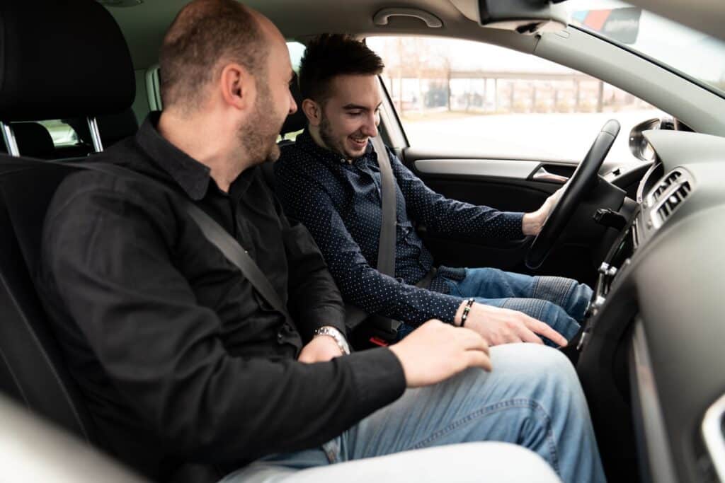 Two men are sitting in a car. The driver, wearing a dark patterned shirt, appears happy and focused on the steering wheel as if recalling his automatic driving lessons in Coventry. The passenger, wearing a black shirt, smiles and looks at the dashboard. Both are wearing seatbelts.