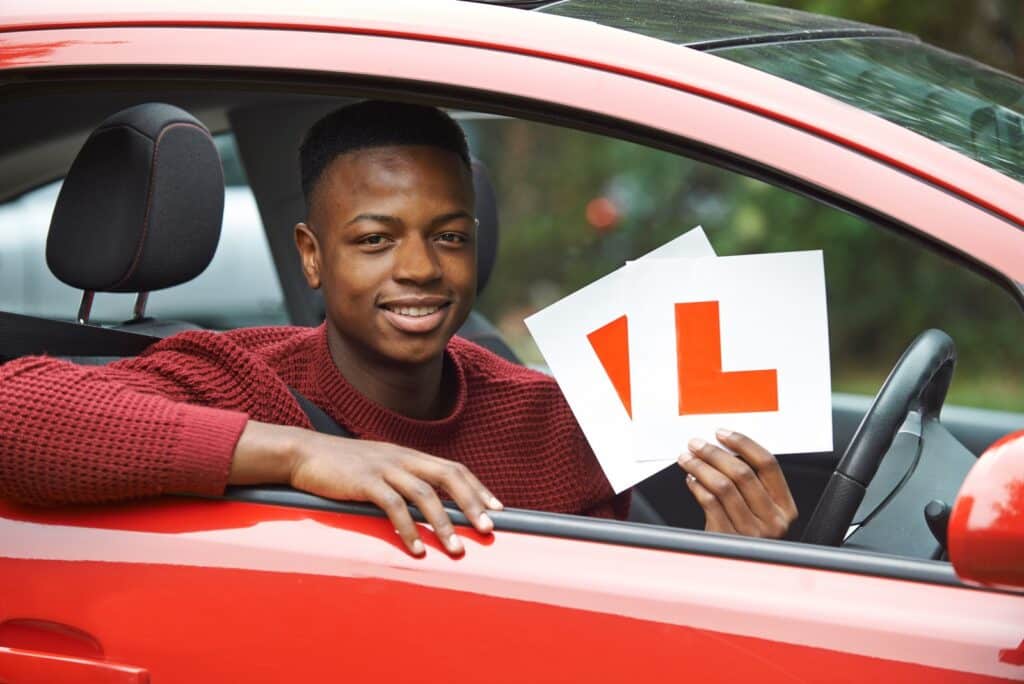 A young man sitting in a red car holds up two large L-plates, commonly used by learner drivers. He is wearing a red sweater and smiling through the open window. The background is a green, blurred outdoor setting.