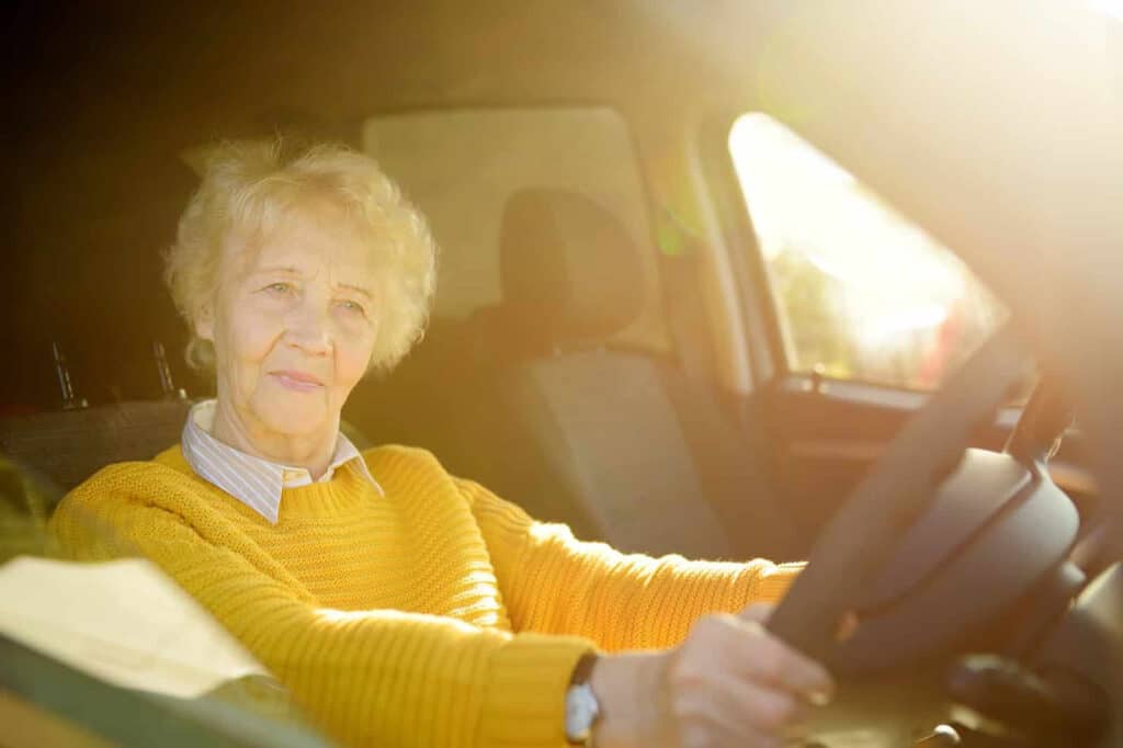 Elderly woman with short white hair wearing a yellow sweater sits in the driver's seat of a car, hands on the steering wheel, with sunlight streaming in through the window.