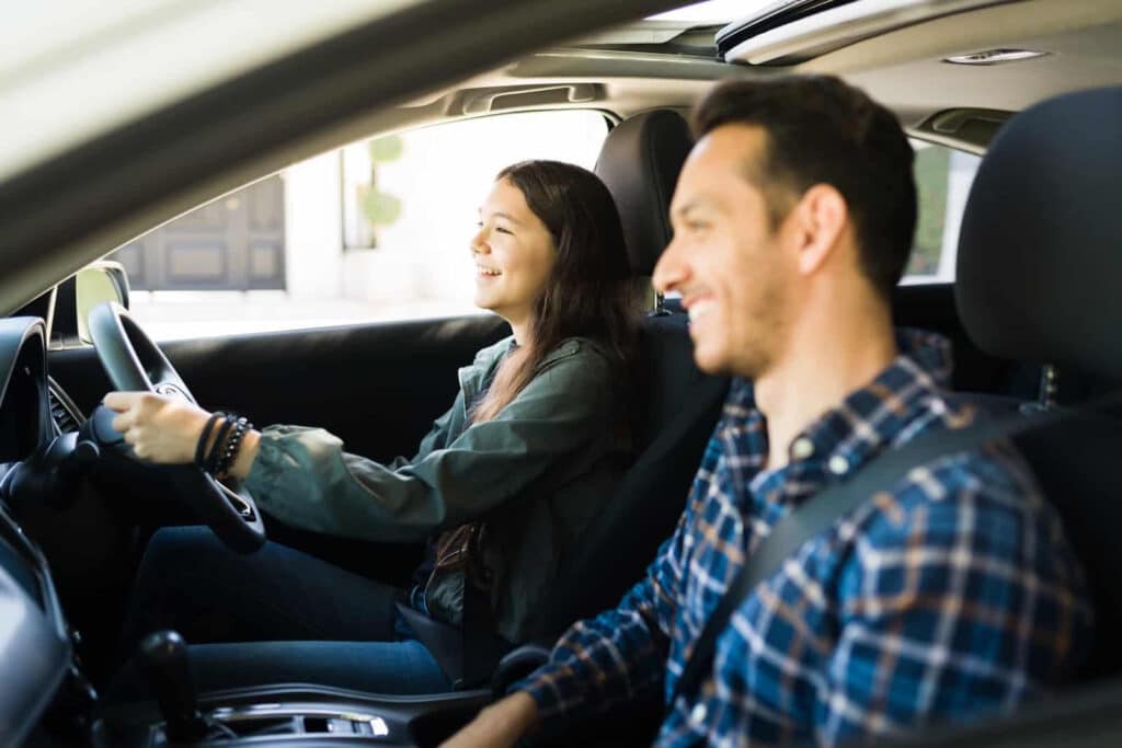 A young woman is smiling while driving a car, with a man in the front passenger seat also smiling. They appear relaxed and content as they look ahead. The photo is taken from the side, capturing the interior of the car and natural light outside.