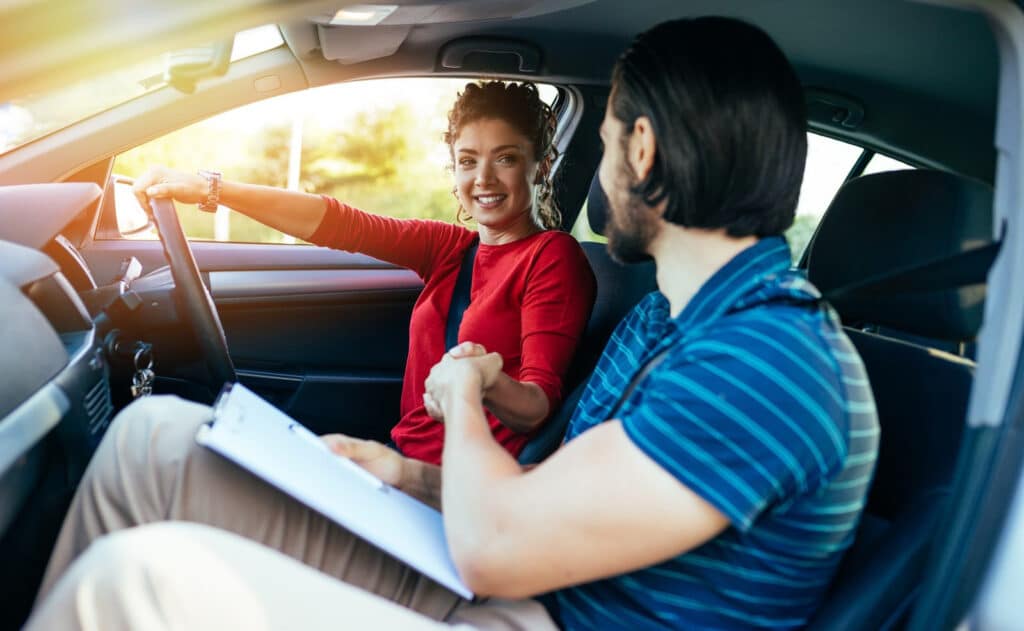 A woman in a red shirt sits in the driver's seat of a car, smiling at a man in a striped shirt holding a clipboard in the passenger seat. They appear to be engaged in conversation, possibly during a driving lesson.