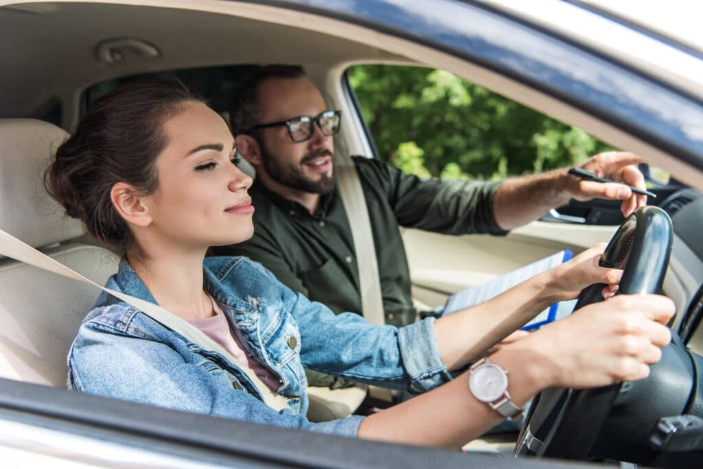 A woman is driving a car while a man in the passenger seat points and talks, possibly instructing her. Both are wearing seatbelts. The interior of the car and greenery outside are visible.