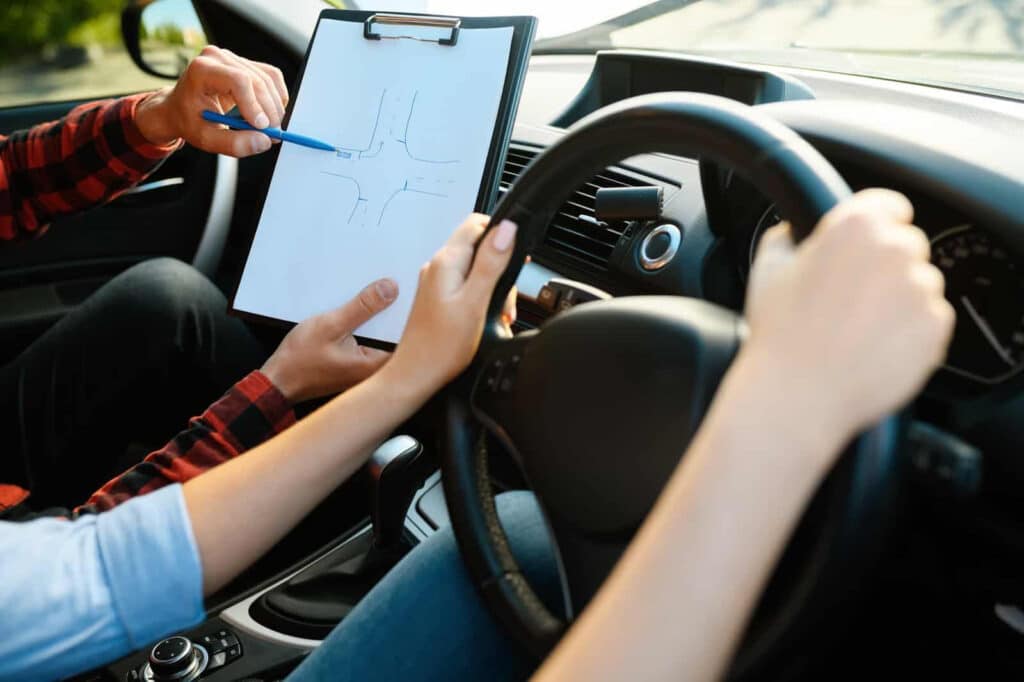 A driving instructor points to a map on a clipboard, guiding a learner who is holding the steering wheel inside a car. Hands of both individuals are visible, emphasizing the instruction process.